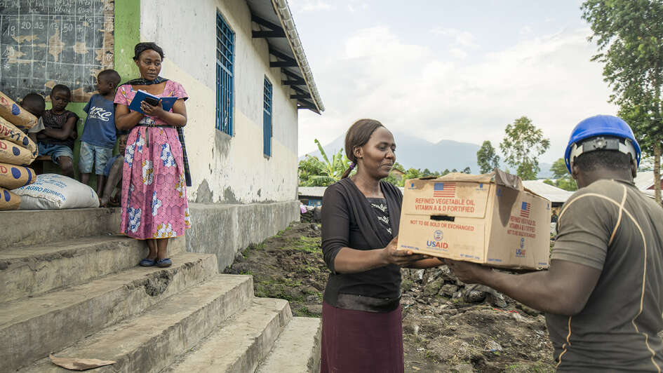 Ein Mann mit blauem Schutzhelm übergibt einer Frau mit schwarzem Shirt und weinrotem Rock vor einem Haus einen Pappkarton, in dem sich - so ist aufgedruckt - Hilfsgüter befinden. Eine Frau in rotem Kleid und mehrere Kinder stehen in einem Hauseingang im Hintergrund und schauen zu.