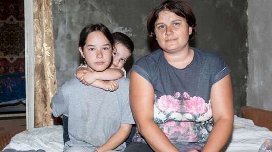 In einer kargen Wohnung sitzen eine Frau und zwei Kinder auf der Kante des Bettes.