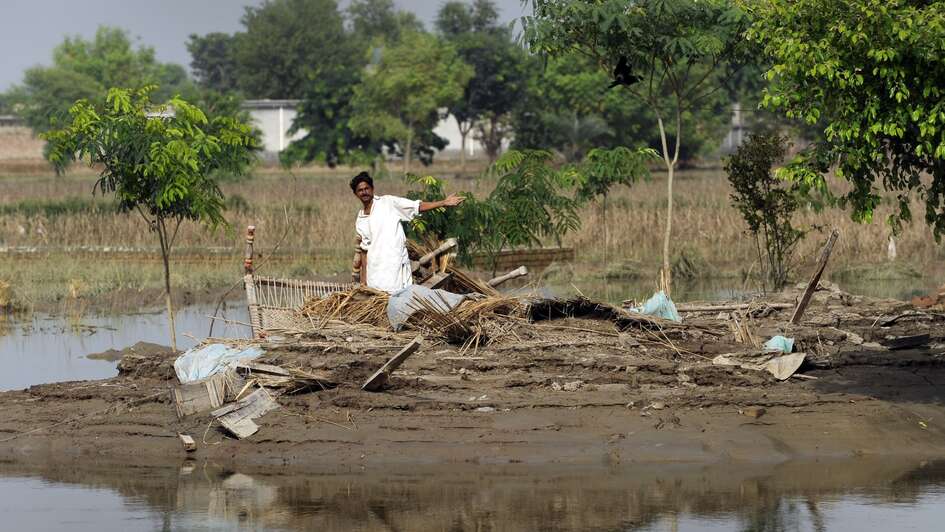 Ein Mann steht auf einem von Wasser umgebenen Stück Land und zeigt auf zerstörte Haushaltsgegenstände.
