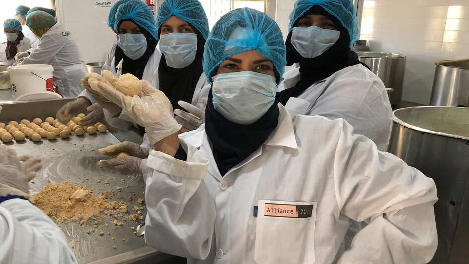 Eine Frau in weißem Kittel, blauer Haarhaube und Mundschutz hält ein Stück Käse hoch.