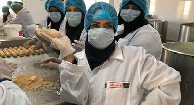 Eine Frau in weißem Kittel, blauer Haarhaube und Mundschutz hält ein Stück Käse hoch.