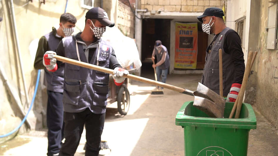 Drei Männer bei der Müllbeseitigung: Ein Mann wirft mit einer großen Schaufel etwas in eine grüne Tonne.