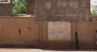 Child standing in front of a house in Sudan after the outbreak of war, smoke rising.