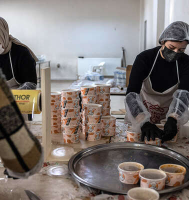 Drei Frauen in Arbeitskleidung verpacken frisch gekochte Mahlzeiten in handliche Transportboxen für die spätere Verteilung.