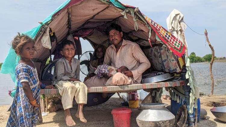 Fluten in Pakistan: Ein Mann, eine Frau, zwei kleine Kinder und ein Baby in ihrer Notunterkunft, die aus Holzstäben, Decken und einer Plastikabdeckung besteht. im Hintergrund ist Wasser zu sehen.