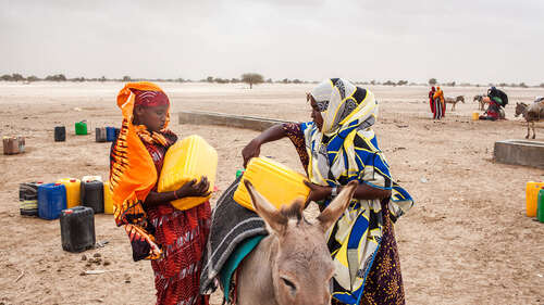 Zwei Frauen heben gelbe Kanister auf einen Esel.