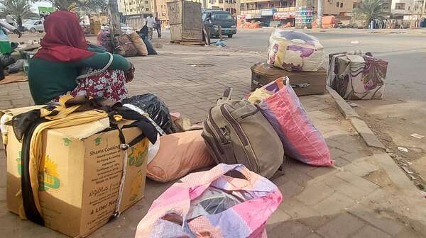 Eine Frau sitzt auf einem Karton auf einer Straße. Um sie herum stehen Taschen.