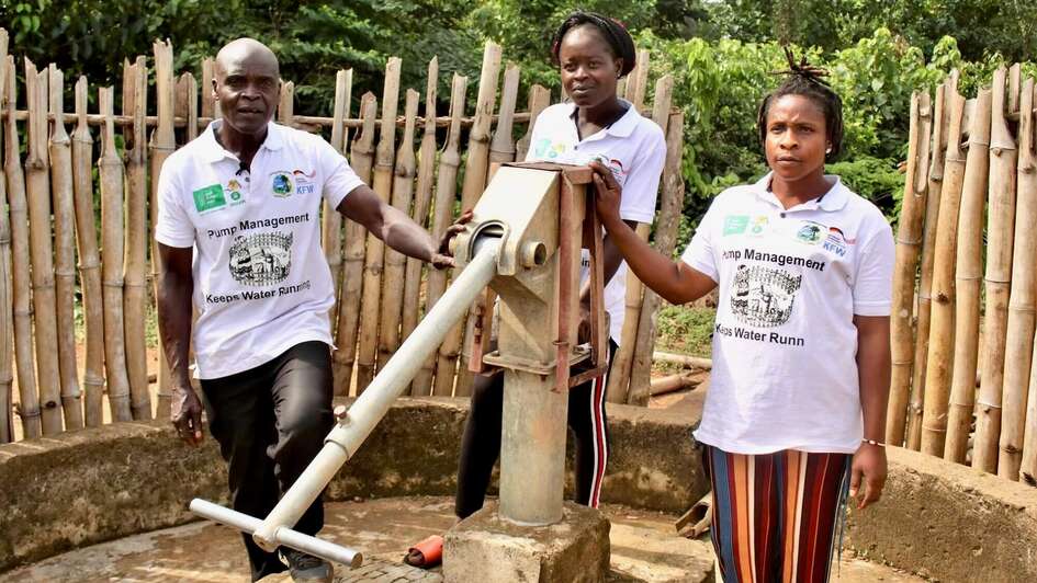 Two women and a man stand next to a water pump and look into the camera.