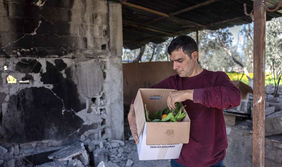 Ein Mann steht vor seinem zerstörten Haus und hält einen Karton mit frisch geerntetem Obst.
