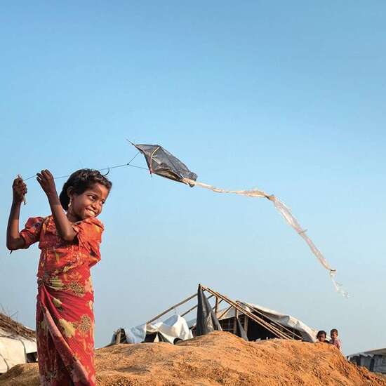 Links steht ein lächelndes Mädchen, das mit einem Drachen spielt. Rechts gehen Menschen vorbei.