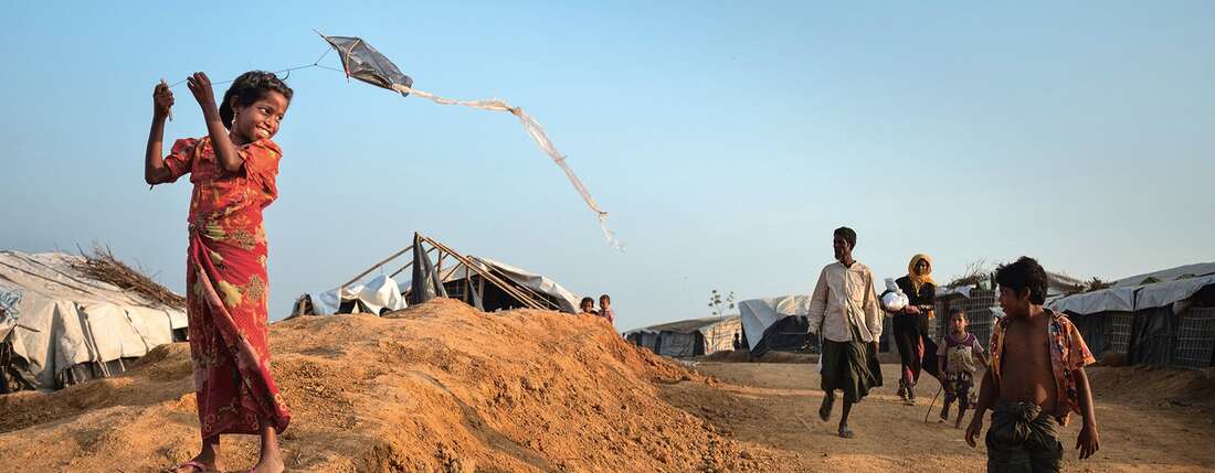 Links steht ein lächelndes Mädchen, das mit einem Drachen spielt. Rechts gehen Menschen vorbei.