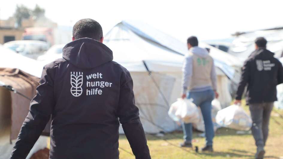 Distribution of hot meals in a refugee camp in Syria. In the foreground, an aid worker with the Welthungerhilfe logo on the back of his jacket.