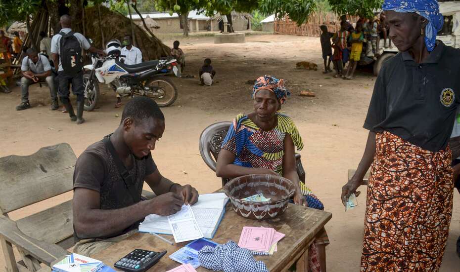 Eine Frau legt Geld in eine Schüssel, die auf einem Tisch steht; ein Mann macht Notizen