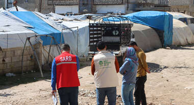 Four people, two of them in the uniforms of aid organizations, in a refugee camp in Syria. The surroundings consist of makeshift shelters covered with blue and white tarpaulins.