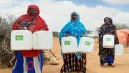 Three women stand side by side, each holding two water containers.